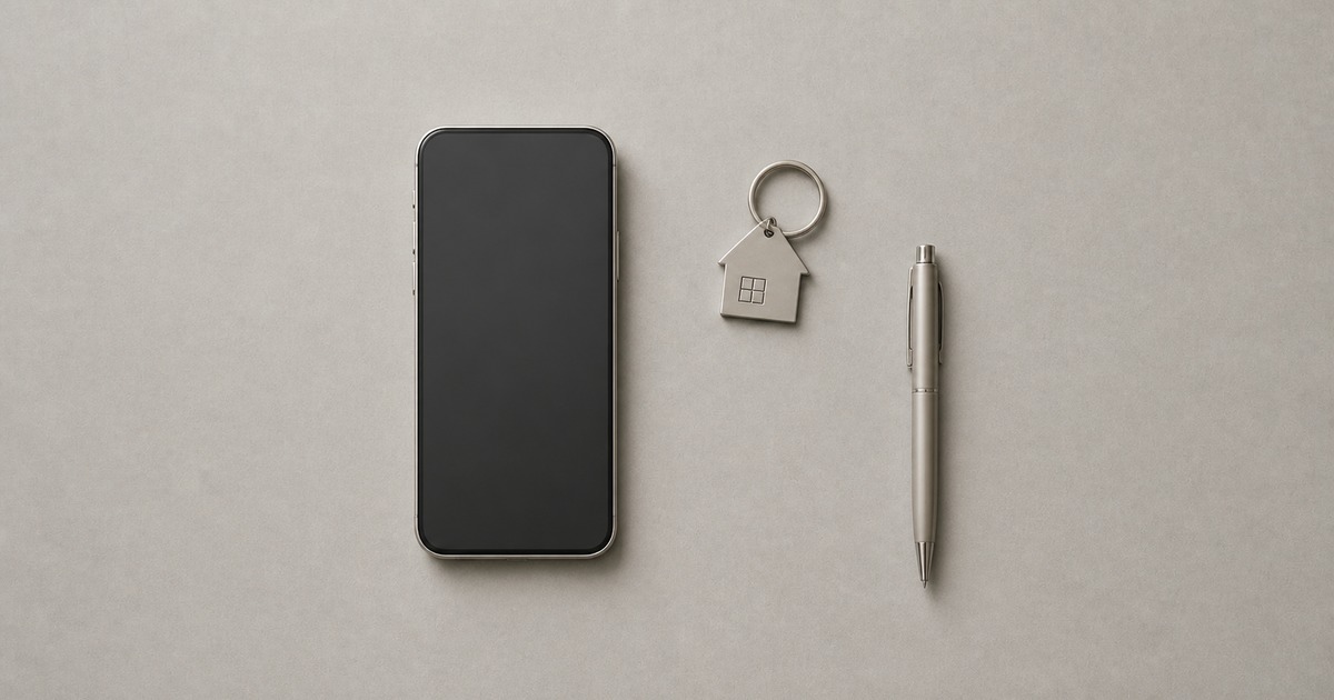 A smartphone, a metallic house-shaped keychain, and a slim pen on a warm grey surface