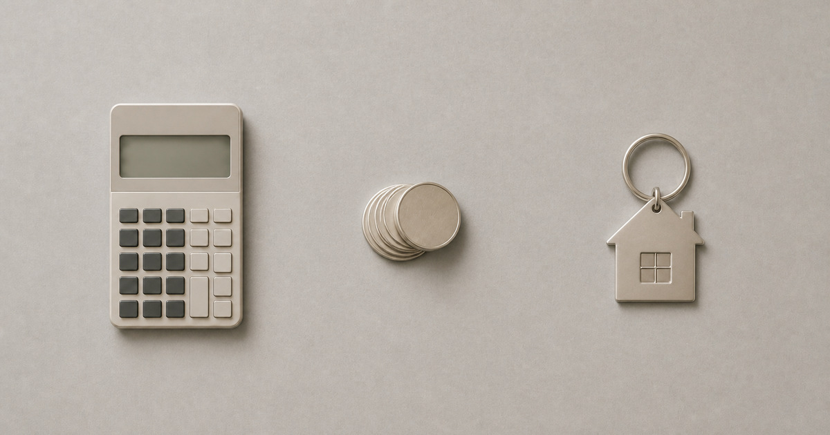 A calculator, a small stack of coins, and a house-shaped keychain on a warm grey surface